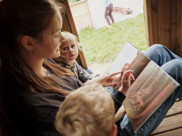 Eine junge Erzieherin in Ausbildung liest zwei Kita-Kindern in einer Garten-Hütte ein Buch vor.