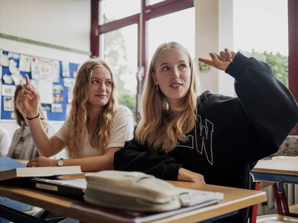 Schülerinnen im Unterricht einer CJD Schule sitzen aufmerksam im Klassenraum, heben motiviert die Hand und beteiligen sich am Unterricht. Im Vordergrund liegen Schulmaterialien auf dem Tisch. Das Bild vermittelt Engagement, moderne Lernatmosphäre und individuelle Förderung an CJD Schulen
