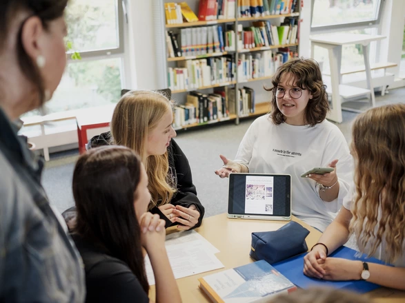 Eine Schülerin präsentiert drei anderen Schülerinnen und einer Lehrerin an einem Tisch sitzend etwas auf einem Tablet. Spenden helfen Bildung in Schulen zu stärken.
