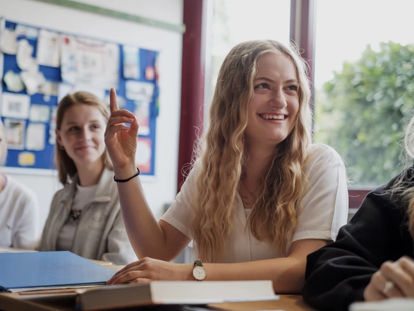 Eine blonde Schülerin sitzt an einem Tisch mit Büchern und Mappen vor sich und meldet sich lächelnd.