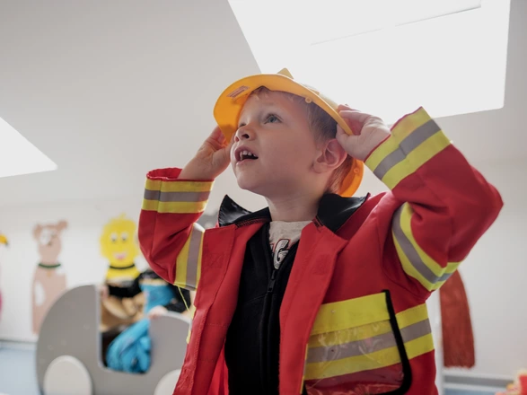 Ein kleiner Junge in einem Feuerwehrkostüm mit roter Jacke und gelbem Helm blickt fasziniert nach oben. Im Hintergrund sind bunte Wanddekorationen mit Tiermotiven zu sehen.