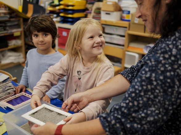Ein Junge und ein Mädchen sind mit einer Erzieherin in einem Bastelraum. Das Mädchen lächelt die Erzieherin fröhlich an. Spenden helfen Kindern Geborgenheit in Kitas zu schenken.