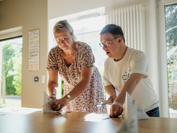 Eine Frau und ein Mann mit Down-Syndrom stehen an einem Tisch und falten gemeinsam Papiere. Beide wirken konzentriert. Sonnenlicht fällt durch das Fenster in den Raum.