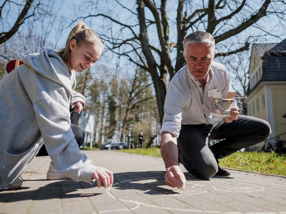 Eine junge Frau und ein älterer Mann malen mit Kreide etwas auf einen Gehweg. 