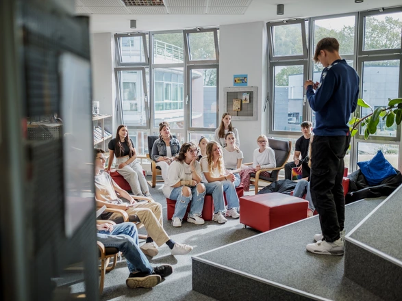 Ein Schüler steht auf einer Art Bühne vor einer kleinen Gruppe von Schülern und Schülerinnen und hält einen Vortrag.