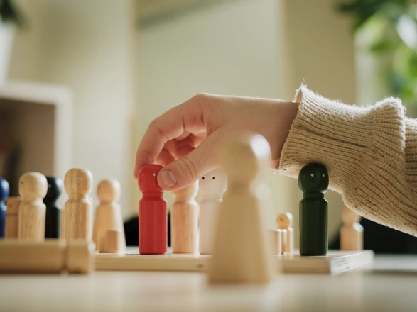 Spielfiguren aus Holz stehen auf einem Holzbrett und einem Tisch. Eine Kinderhand ist dabei eine der Spielfiguren zu versetzen.