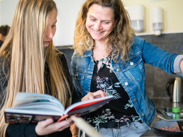 Eine Jugendliche steht neben einer Pädagogin am Herd. Sie schauen beide in ein Kochbuch.