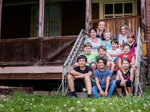  Eine Kindergruppe sitzt mit drei Pädagoginnen auf einer Treppe vor einem alten Gebäude.