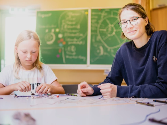 Ein Kind und eine Erwachsene sitzen am Tisch. Vor ihnen liegen verschiedene Bastelmaterialien, z.B. Schnüre und Perlen.