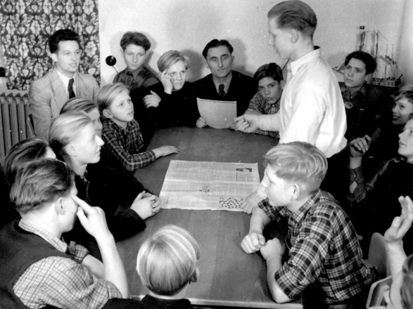 Schwarz-weiß-Fotografie einer größeren Gruppe Jugendlicher und junger Männer, die in den 1950er Jahren um einen Tisch versammelt sind. Ein Junge steht auf und spricht, während die anderen aufmerksam zuhören. Auf dem Tisch liegt eine aufgeschlagene Zeitung. Die Szene wirkt wie eine demokratische Gesprächsrunde oder ein Mitbestimmungsgremium in einer pädagogischen Einrichtung.