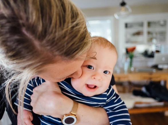 Eine erwachsene Person hält ein Baby im Arm und küsst es liebevoll auf die Wange. Das Baby trägt ein blau-weiß gestreiftes Shirt und lächelt. Im Hintergrund ist ein heller, wohnlicher Raum mit Tisch, Stühlen und Blumen zu sehen.