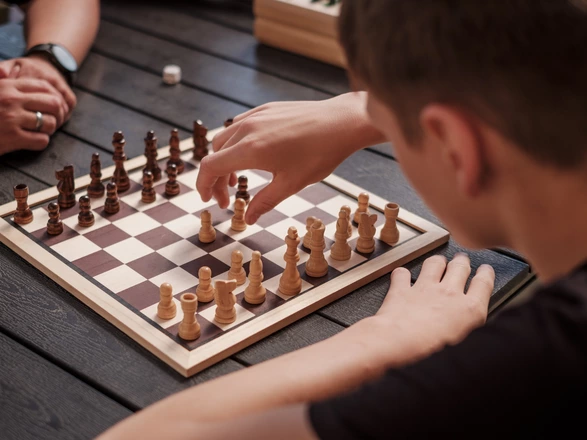 Ein Junge sitzt an einem Tisch und spielt Schach. Er greift mit der rechten Hand nach einer Figur auf dem Schachbrett. Gegenüber sind die Hände seines Spielpartners zu sehen.