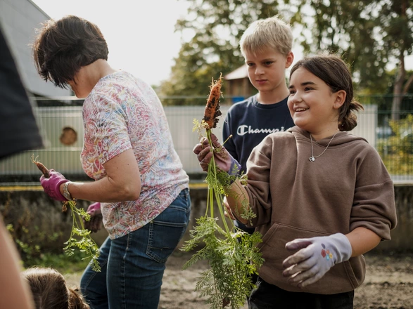 Ein Mädchen, ein Junge und eine Lehrerin sind in einem Schulgarten. Das Mädchen hält eine frisch geerntete Karotte hoch. Spenden stärken Bildungsangebote in unseren CJD Schulen.