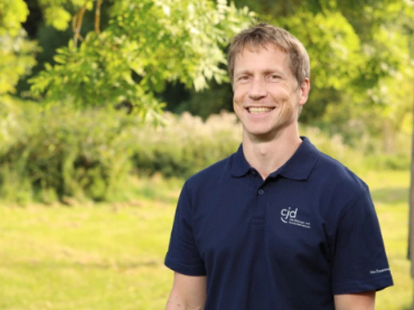 Ein Mann mit kurzen, hellbraunen Haaren steht lächelnd im Freien vor grüner, sommerlicher Naturkulisse. Er trägt ein dunkelblaues Poloshirt mit dem weißen CJD-Logo und dem Schriftzug „Das Bildungs- und Sozialunternehmen“. Im Hintergrund sind Bäume, Wiese und unscharfe Pflanzen zu sehen, die eine freundliche und natürliche Atmosphäre schaffen.