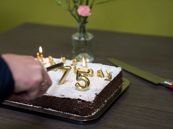 Ein rechteckiger Schokoladenkuchen mit Puderzucker ist mit goldenen Kerzen in Form der Zahl „75“ und weiteren Buchstaben dekoriert. Eine Hand entzündet die Kerzen mit einem Stabfeuerzeug. Daneben liegt ein großes Messer mit rotem Griff auf dem Tisch. Im Hintergrund steht eine kleine Glasvase mit einer rosa Blume vor grünlicher Wand.