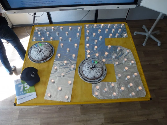 Auf einem gelben Tisch ist die Zahl 75 mit Alufolie ausgelegt und von vielen kleinen Teelichtern umrahmt. Zwei Kuchen mit Geburtstagskerzen in Form einer „7“ und „5“ stehen auf der Fläche. Daneben liegen eine schwarze CJD-Kappe und ein Flyer mit der Aufschrift „75 Jahre“. Die Dekoration erinnert an eine Feier zum Jubiläum. Im Hintergrund sind Teile eines Bildschirms und Möbel zu erkennen.