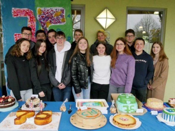 Eine Gruppe Jugendlicher steht dicht zusammen hinter einem Tisch mit vielen selbstgebackenen Kuchen. Auf dem Tisch liegen bunt verzierte Torten, darunter eine mit der Aufschrift „75“ und eine mit dem CJD-Logo. Im Hintergrund hängt ein farbig gestaltetes Plakat mit einer großen „75“. Alle lächeln in die Kamera, die Szene wirkt fröhlich und festlich.