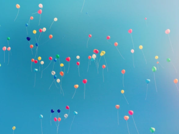 Vor strahlend blauem Himmel steigen zahlreiche bunte Luftballons in die Höhe. Die Ballons sind in verschiedenen Farben – Rot, Gelb, Blau, Grün, Rosa und Orange – und mit langen weißen Schnüren versehen. Sie schweben frei verteilt im Bild und erzeugen eine fröhliche, leichte und festliche Stimmung.