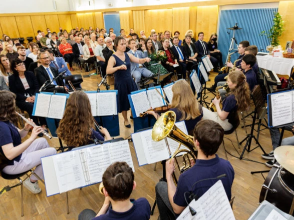 Eine Schülerband mit Blasinstrumenten und Schlagzeug spielt in einem Saal vor großem Publikum. Die Dirigentin steht in der Mitte und leitet das Ensemble. Im Hintergrund sitzen zahlreiche Gäste in feierlicher Kleidung, aufmerksam den jungen Musikerinnen und Musikern lauschend. Die Szene vermittelt eine festliche, lebendige Atmosphäre voller Musik und Gemeinschaft.
