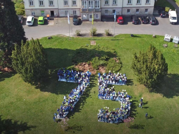 Aus der Luftperspektive ist eine große Menschenmenge vor einem historischen Gebäude zu sehen, die die Zahl „75“ formt. Viele Teilnehmende tragen bunte Luftballons in Blau, Grün und Gelb, wodurch die Jubiläumszahl eindrucksvoll hervorgehoben wird. Die Aktion auf der Rasenfläche markiert das 75-jährige Bestehen des CJD und symbolisiert Gemeinschaft, Zusammenhalt und Feierlichkeit.