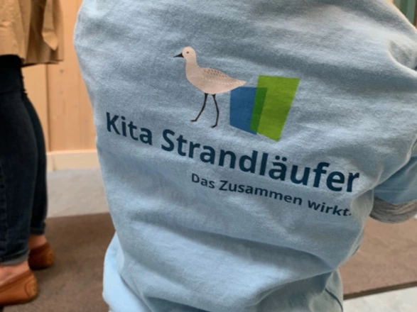 Ein kleines Kind trägt ein hellblaues T-Shirt mit dem Aufdruck „Kita Strandläufer – Das Zusammen wirkt.“ sowie einer Möwe als Logo. Das Kind steht mit dem Rücken zur Kamera, wodurch der Schriftzug und das Logo gut sichtbar sind. Im Hintergrund sind Erwachsene im Eingangsbereich der Einrichtung erkennbar. Die Szene vermittelt den Eindruck von Zugehörigkeit und Gemeinschaft in der Kindertagesstätte.