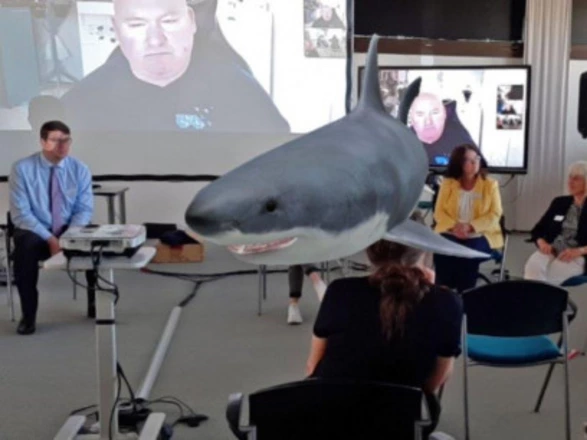 Eine Gruppe von Menschen sitzt in einem Seminarraum, während auf einem Bildschirm im Hintergrund eine Videokonferenz läuft. Im Vordergrund ist ein großer Hai als digitale 3D-Projektion zu sehen, der mitten durch den Raum zu schwimmen scheint. Die Szene zeigt den Einsatz moderner Augmented-Reality-Technologie, die das Treffen interaktiv und visuell eindrucksvoll gestaltet