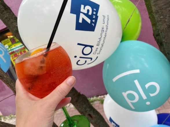 Eine Hand hält ein Glas mit einem Aperitif-Getränk, garniert mit Orangenscheiben und Strohhalm. Im Hintergrund sind bunte Luftballons mit dem CJD-Logo und dem Schriftzug „75 Jahre“ zu sehen. Die Szene vermittelt sommerliche Feierlaune und markiert den Anlass des 75-jährigen Jubiläums des CJD.
