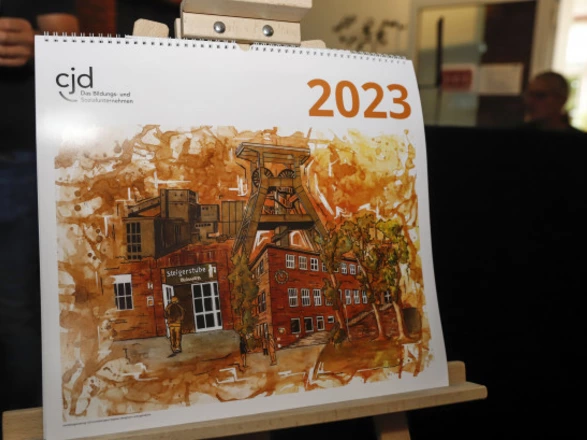 Ein Kalender des CJD für das Jahr 2023 steht auf einer Staffelei. Auf dem Titelblatt ist eine künstlerische Illustration eines Bergwerksgebäudes mit Fördertürmen und rotem Backstein zu sehen. Oben links steht das CJD-Logo mit dem Schriftzug „Das Bildungs- und Sozialunternehmen“, oben rechts die Jahreszahl „2023“ in Orange.