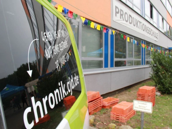 Ein grünes Auto mit der Aufschrift „chronik.cjd.de – Gehen Sie mit uns auf Entdeckungsreise. 75 Jahre CJD“ steht vor einem Gebäude der Produktionsschule. Am Gebäude hängt bunte Wimpel-Dekoration, und auf dem Hof sind gestapelte rote Kunststoffkisten zu sehen.