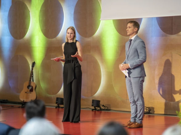 Eine Frau im schwarzen Outfit und ein Mann im hellgrauen Anzug moderieren eine Veranstaltung auf einer Bühne mit rotem Boden. Im Hintergrund ist eine Wand mit runden Ausfräsungen, die farbig beleuchtet wird. Neben der Bühne steht eine Akustikgitarre. Beide Personen sprechen vor einem Publikum, das im Vordergrund unscharf zu sehen ist.