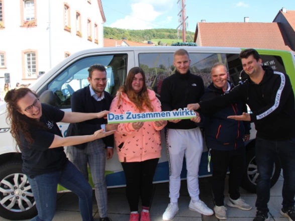 Sechs Personen stehen lachend vor einem CJD-Bus und halten gemeinsam ein Schild mit der Aufschrift „Das Zusammen wirkt.“ in die Kamera. Die Gruppe besteht aus jungen Erwachsenen und Mitarbeitenden, die eng nebeneinanderstehen und den Teamgeist symbolisieren. Im Hintergrund sind Häuser und ein sonniger Himmel zu sehen.