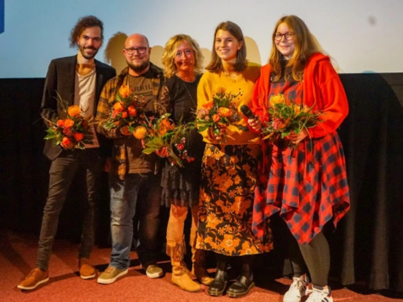 Fünf Personen stehen nebeneinander auf einer Bühne vor einer Leinwand und halten Blumensträuße in den Händen. Sie lächeln in die Kamera, eine Person trägt zusätzlich ein Mikrofon. Die Gruppe wirkt feierlich und harmonisch, als sei es der Abschluss oder die Ehrung bei einer Veranstaltung, etwa einer Filmpremiere oder Preisverleihung.