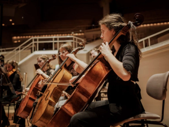 Auf dem Foto ist eine Cellistin zu sehen, die konzentriert während eines Orchestereinsatzes spielt. Mehrere Musikerinnen und Musiker sitzen nebeneinander, ebenfalls mit Celli, in einem Konzertsaal mit heller Bühne und moderner Architektur. Die Stimmung wirkt konzentriert, feierlich und professionell – ganz so, wie man es bei einem großen Orchesterkonzert erwarten würde.