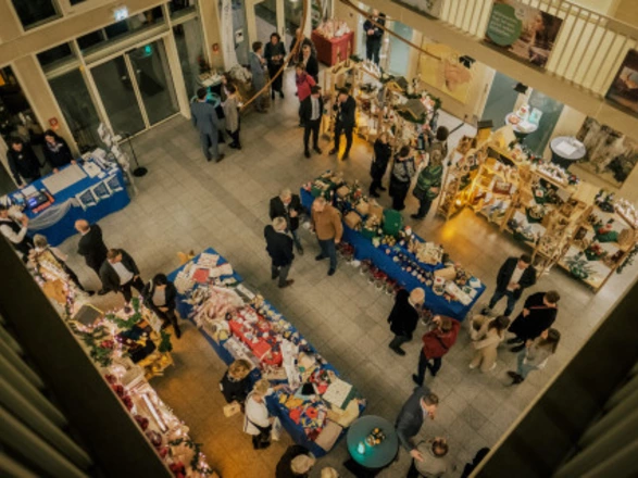 Blick von oben auf einen weihnachtlich dekorierten Markt in einer Halle. Menschen schlendern zwischen Ständen mit bunten Handarbeiten, Dekorationen und Geschenkartikeln. Einige Besucher unterhalten sich, andere betrachten die Auslagen. Festliche Beleuchtung und geschmückte Tische sorgen für eine warme, gemütliche Atmosphäre.