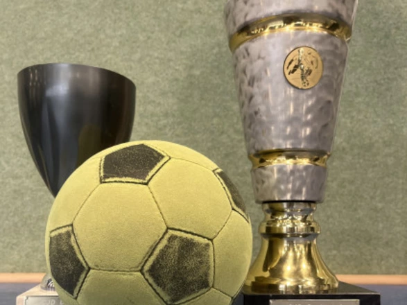 Ein gelb-grüner Softfußball liegt vor zwei Pokalen. Der rechte Pokal ist groß, silbern mit goldenen Akzenten und trägt die Gravur „CJD Dortmund Soccercup Wanderpokal“. Links daneben steht ein kleinerer, dunkler Pokal. Das Bild vermittelt eine sportliche Atmosphäre und erinnert an ein Fußballturnier mit Siegerauszeichnung.