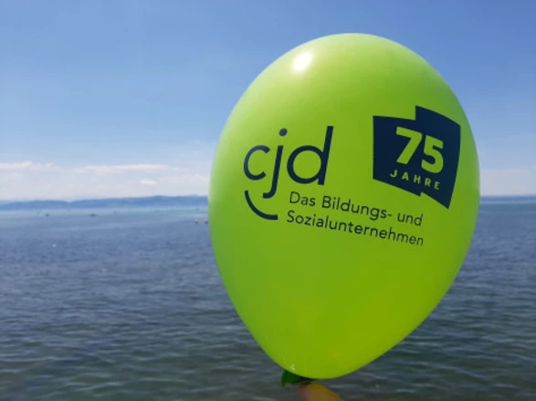 Ein grüner Luftballon mit dem blauen Aufdruck „cjd – Das Bildungs- und Sozialunternehmen“ sowie dem Logo „75 Jahre“ schwebt vor blauem Himmel am Ufer eines Sees. Das Bild symbolisiert das Jubiläum des CJD in festlicher, sommerlicher Atmosphäre.