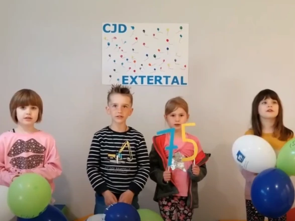 Vier Kinder stehen nebeneinander vor einer Wand mit einem bunten Plakat „CJD Extertal“. Sie halten Luftballons in Blau, Grün und Weiß mit CJD-Logo sowie eine bunte „75“-Zahl in den Händen. Die Szene vermittelt Freude und Zusammenhalt und zeigt eine Aktion zum 75-jährigen Jubiläum des CJD.