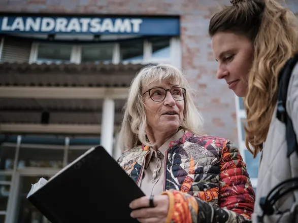 Zwei Frauen im Gespräch vor dem Landratsamt: Eine ältere Frau mit Brille und bunter Jacke hält eine schwarze Mappe in der Hand und spricht mit einer jüngeren Frau. Das Bild symbolisiert Beratung, Unterstützung und Behördenkontakt im öffentlichen Raum.