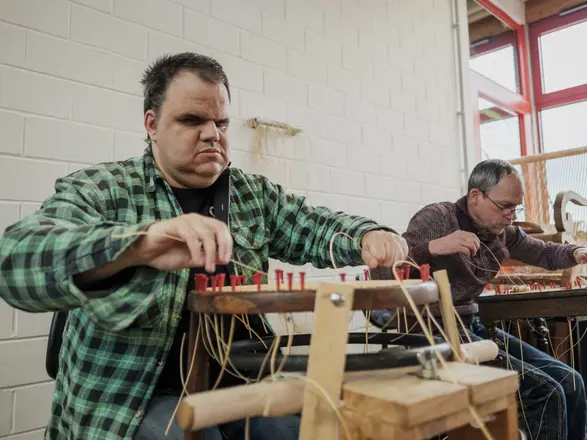 Zwei Männer arbeiten konzentriert an der Herstellung oder Reparatur von Stühlen in einer Werkstatt. Im Vordergrund sitzt ein Mann mit grün-karierter Jacke, der handwerklich mit Naturmaterialien arbeitet. Das Bild zeigt inklusive Beschäftigung, handwerkliches Geschick und Teilhabe am Arbeitsleben im CJD.
