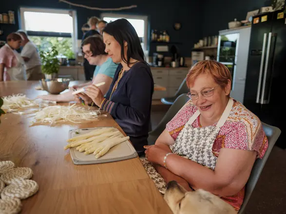 Mehrere Menschen schälen gemeinsam Spargel an einem großen Küchentisch, während eine Frau im Vordergrund lächelnd einen Hund streichelt. Die Szene vermittelt Inklusion, gemeinschaftliches Kochen und Freude am Miteinander im CJD