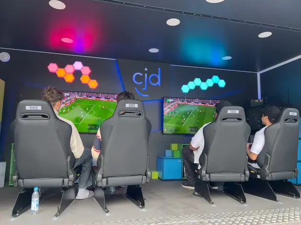 Einblick in den Game4All-Anhänger. 4 Personen sitzen auf 4 Gaming-Stühlen. Auf zwei Monitoren ist ein FIFA-Spiel zu sehen.