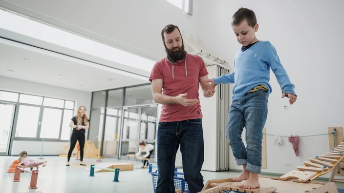 Ein Erzieher hilft einem kleinen Jungen über eine Holz-Brücken in einem Gymnastikraum einer CJD Kita.