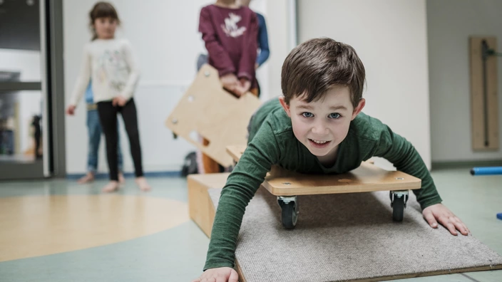 Junge liegt lachend auf einem Rollbrett. Im Hintergrund warten weitere Kinder auf ihren Einsatz. Das Bild vermittelt spielerische Bewegung, Gemeinschaft und Freude an inklusiver Förderung im CJD.