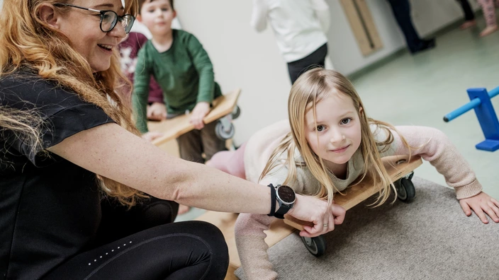 Ein blondes Mädchen liegt auf einem Rollbrett in einem Sportraum und wird dabei von einer Erzieherin unterstützt. Im Hintergrund sind weitere Kinder zu sehen. Spenden helfen besondere Momente in Kitas zu schaffen.