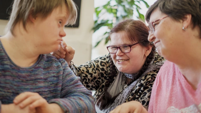 Drei Frauen mit und ohne Behinderung sitzen gemeinsam am Tisch, lachen und tauschen sich aus. Bei uns stehen Miteinander, Wertschätzung und echte Teilhabe im Mittelpunkt. Erlebe die Vielfalt unserer inklusiven Angebote – jetzt informieren und Gemeinschaft erleben!