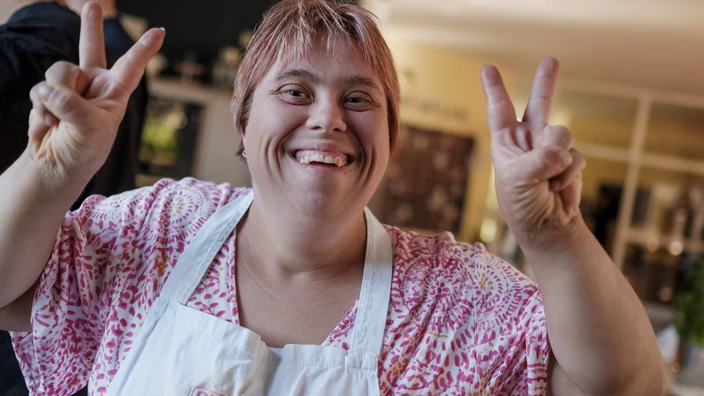 Eine Frau mit Kochschürze lächelt in die Kamera und zeigt mit ihren Fingern das Peace Zeichen. Spenden ermöglichen Teilhabe für Menschen mit Behinderungen.