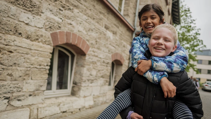 Ein blondes Mädchen trägt ein braunhaariges Mädchen huckepack draußen neben einem Gebäude. Beide lachen dabei. Durch Spenden kann das CJD die Bildung in ihren Schulen stärken.  