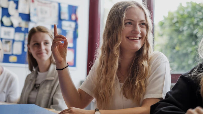 Eine blonde Schülerin sitzt an einem Tisch mit Büchern und Mappen vor sich und meldet sich lächelnd.