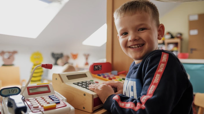 Ein lächelnder Junge spielt mit einer Kasse. Spenden helfen Kindern Geborgenheit in Kitas zu schenken.