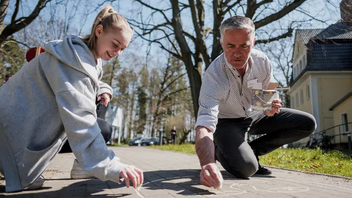 Eine junge Frau und ein älterer Mann malen mit Kreide etwas auf einen Gehweg. 